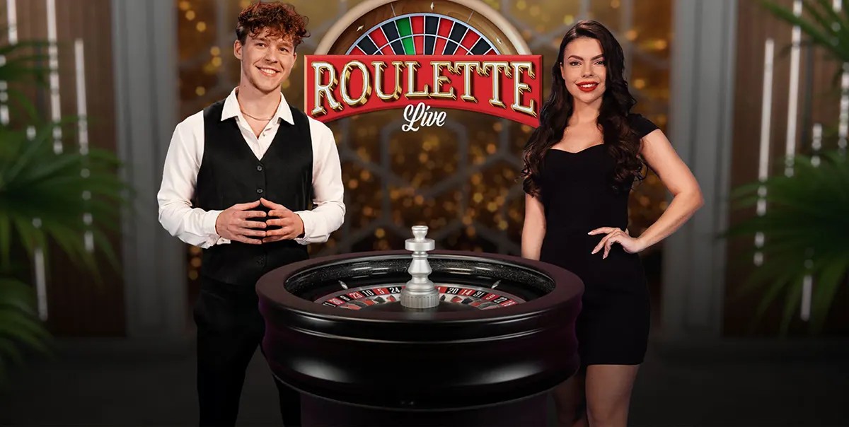 Two live roulette hosts—a man in a black vest and a woman in a black dress—stand beside a roulette wheel under a “Roulette Live” sign in a glossy casino-style studio.